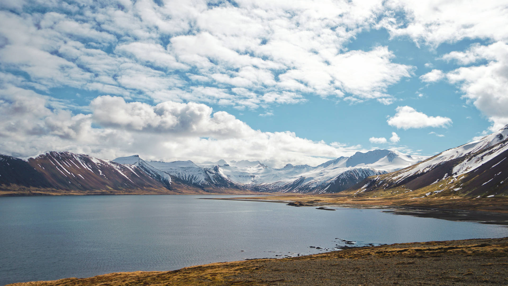 Fjellinnsjø med snødekte fjell i det fjerne og en skyet himmel.