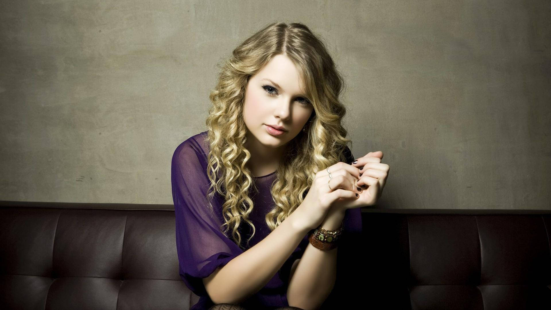 Sittende krøllete hår Taylor Swift i lilla mesh-topp bakgrunnsbilde.