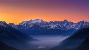 En fantastisk ultravid utsikt over snødekte fjelltopper ved soloppgang med tåkete dal.